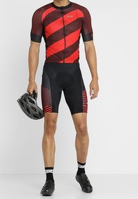Jersey de ciclismo rojo y negro con rayas diagonales, con cremallera y textura de malla. Shorts negros con acentos a rayas. Casco en la mano.