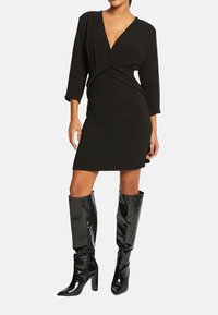 Robe noire portefeuille à manches longues et décolleté en V profond, associée à des bottes en cuir verni hauteur genou avec une finition texturée.
