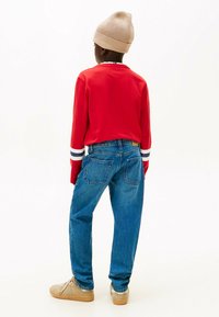 Camisa roja de manga larga con rayas azul marino y blancas en los puños, jeans de mezclilla azules, gorro de punto beige y zapatillas beige. Vista trasera.