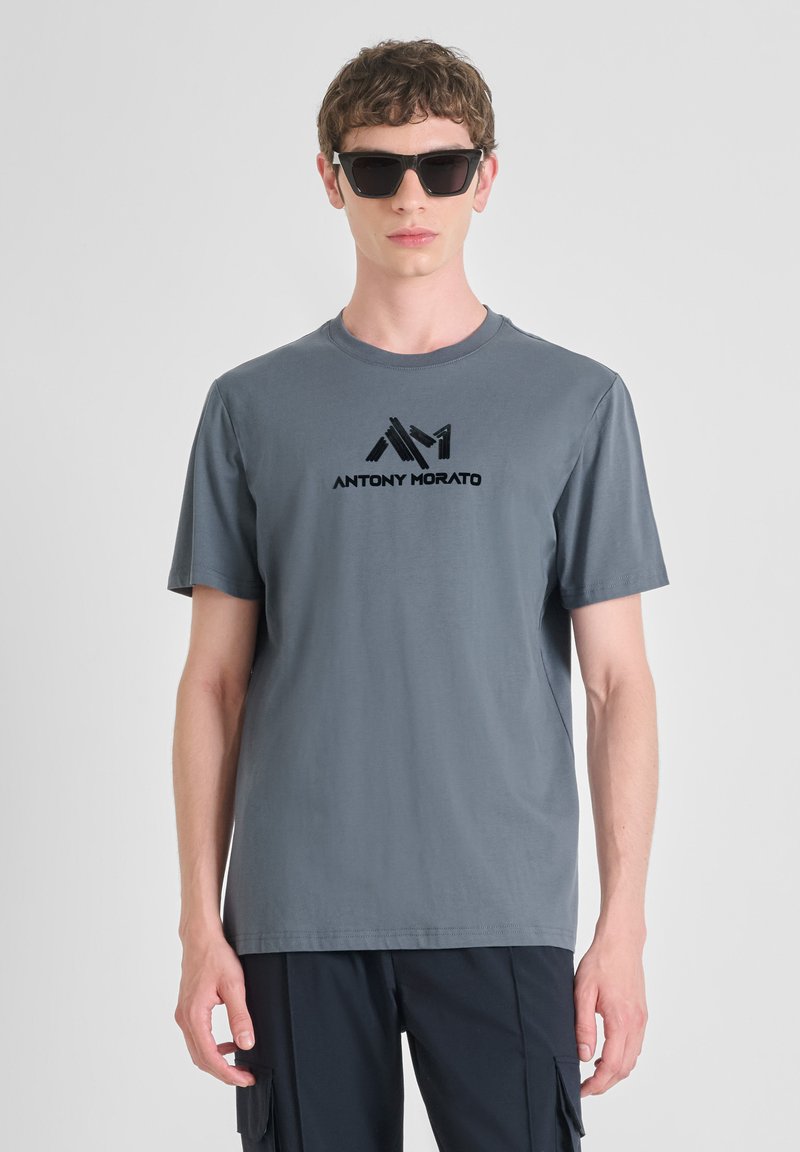 Siva pamukasta majica s prednjim logotipom u crnoj boji, okruglim izrezom i kratkim rukavima, u kombinaciji s crnim naočalama.