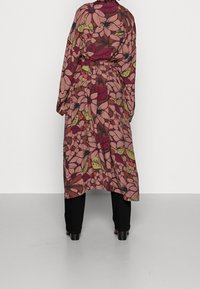 Bloemen maxi-jurk in roze, rode en groene tinten, met een taillering, lange mouwen en een vloeiende achterkant, gecombineerd met zwarte broek.