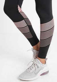 Czarne legginsy z różowymi poziomymi paskami i teksturowanym wykończeniem, zestawione z jasnoszarymi butami sportowymi z siateczkowym cholewką i poduszką powietrzną.