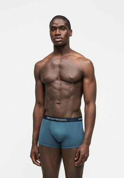Muskularny mężczyzna stoi, ubrany w niebieskie bokserki Emporio Armani, na jednolitym białym tle, stoi twarzą do przodu z neutralnym wyrazem twarzy.
