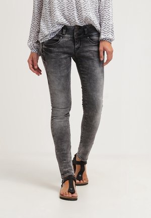 Person iført falmede sorte skinny jeans, mønstret langærmet top og sorte åbne sandaler, der går på en lys baggrund.