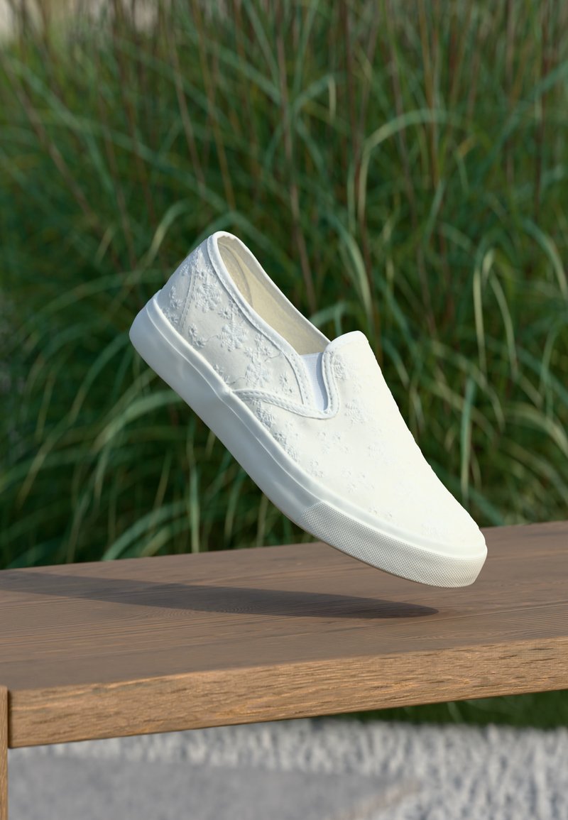 Λευκό αθλητικό παπούτσι χωρίς κορδόνια με κεντήματα λουλουδιών, αιωρείται πάνω από ξύλινο πάγκο, με πράσινα φύλλα στο φόντο.