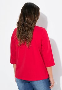 Rotes Oberteil mit drei Viertelärmeln, aus einem glatten Stoff gefertigt. Mit einem runden Ausschnitt und einem lockeren Schnitt. Getragen mit blauen Jeans.