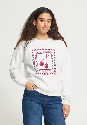 Mujer con cabello largo y rizado que lleva una sudadera blanca con patrón de cerezas rojas y corazones, vaqueros azules, mano en el bolsillo, fondo neutro.