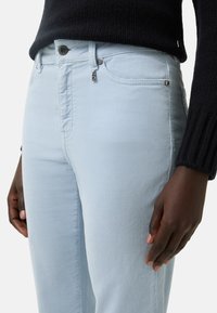 Zblízka osoba v světle modrých kordových kalhotách a tmavém žebrovaném svetru, s uvolněnýma rukama podél těla.