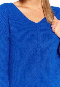 Blå stickad tröja med V-ringning och texturerad ribbning. Har ett centralt kabelmönster som löper ner fram och långa ärmar.