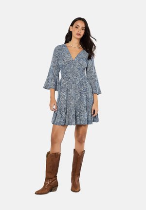 Blaues Paisley-Kleid mit langen Glockenärmeln, einem V-Ausschnitt und einem schwingenden Rock. Kombiniert mit braunen Cowboy-Stiefeln und einer Perlenkette.