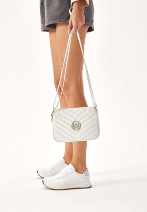Borsa a tracolla bianca imbottita con logo dorato, forma rettangolare, chiusura con zip e tracolla lunga, abbinata a sneaker bianche su uno sfondo neutro.