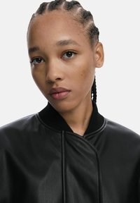 Veste bomber en cuir noir avec col côtelé, texture lisse et détails de coutures à l'avant. Le modèle présente une coiffure tressée et un maquillage naturel.