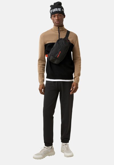 Sweater i sort og tan med farveblok, sort crossbody taske med orange tekst, sorte bukser og lyse sneakers. Modellen bærer en sort beanie med hvid tekst.