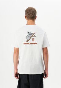 Person med hvit T-skjorte med grafikk av en koala på en gren og teksten "Rip Curl Australia Road trippin' in the high country" på ryggen.
