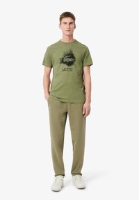 Oliwkowy bawełniany T-shirt z czarnym logo Lacoste i nadrukiem graficznym, w połączeniu z pasującymi oliwkowymi dresami i białymi trampkami.