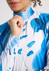 Långärmad atletisk topp i vitt med blå abstrakta mönster. Design med halv ståkrage och dragkedja samt åtsittande ärmslut i ett mjukt, lätt material.