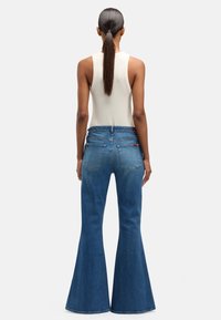 Dżinsy o wysokim stanie, rozszerzane, w kolorze niebieskim, z miękką teksturą denimu, widocznym przeszyciem i detalami szwów. Widok od tyłu ukazuje dwie tylne kieszenie.