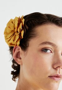 Un giovane con la pelle chiara e i capelli corti castani indossa un grande fiore di stoffa giallo fissato sopra l'orecchio sinistro su sfondo bianco.