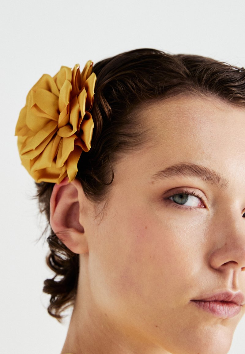 Un giovane con la pelle chiara e i capelli corti castani indossa un grande fiore di stoffa giallo fissato sopra l'orecchio sinistro su sfondo bianco.