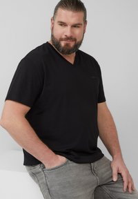 Czarna koszulka z dekoltem w serek z miękkiej bawełny, z krótkimi rękawami i luźnym krojem. Model nosi szare dżinsy i stoi z rękami w kieszeniach.