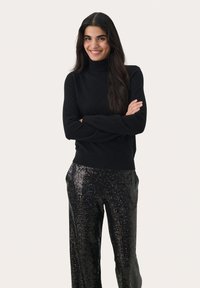 Melnās turtlenecks kreps ar spīdīgām melnām sequin platām biksēm, izceļot gludu tekstūru un pieguļošu siluetu.