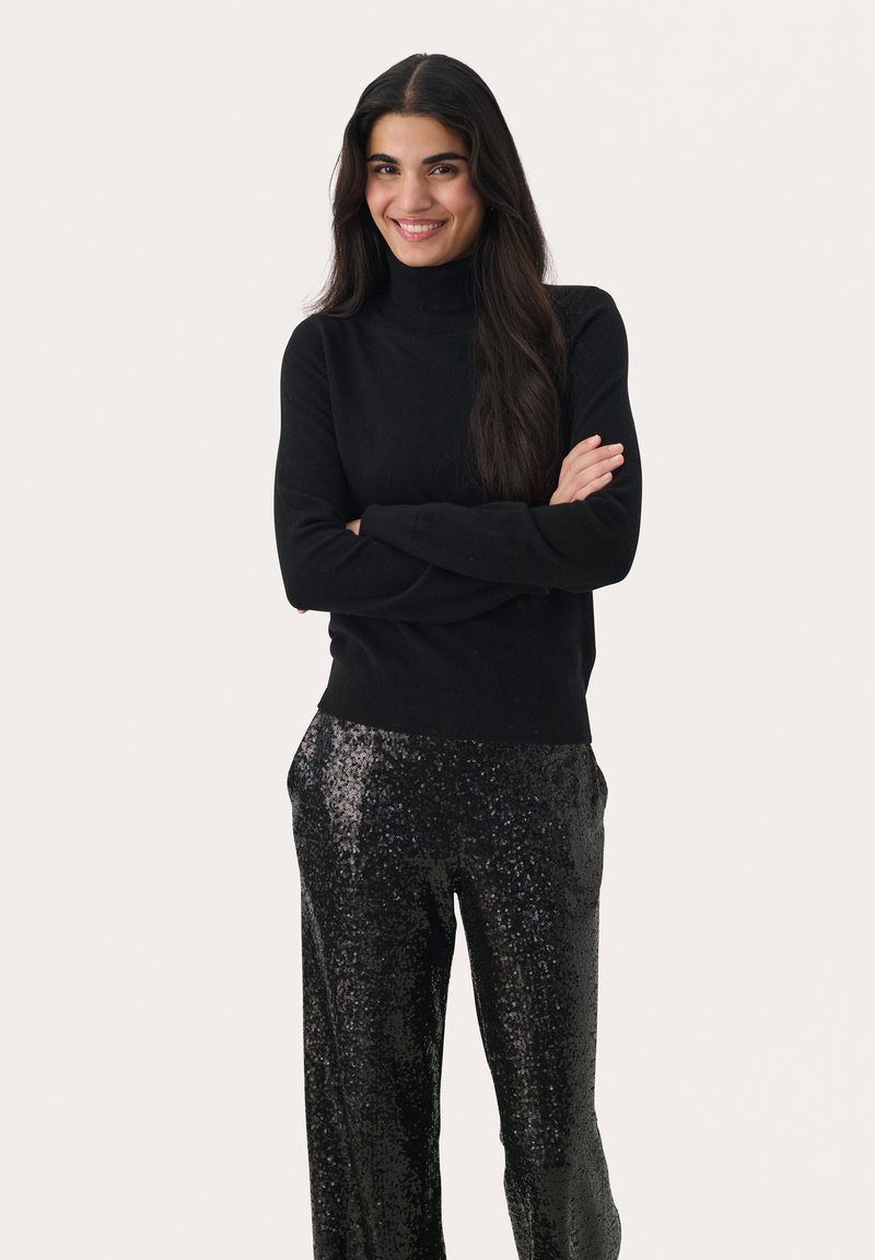 Melnās turtlenecks kreps ar spīdīgām melnām sequin platām biksēm, izceļot gludu tekstūru un pieguļošu siluetu.