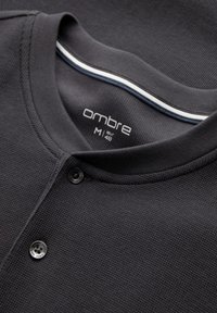 Nahaufnahme eines dunkelgrauen Waffelstrick-Henley-Shirts mit zwei Knöpfen und kontrastierenden Details am inneren Kragen in Weiß und Blau gestreift.