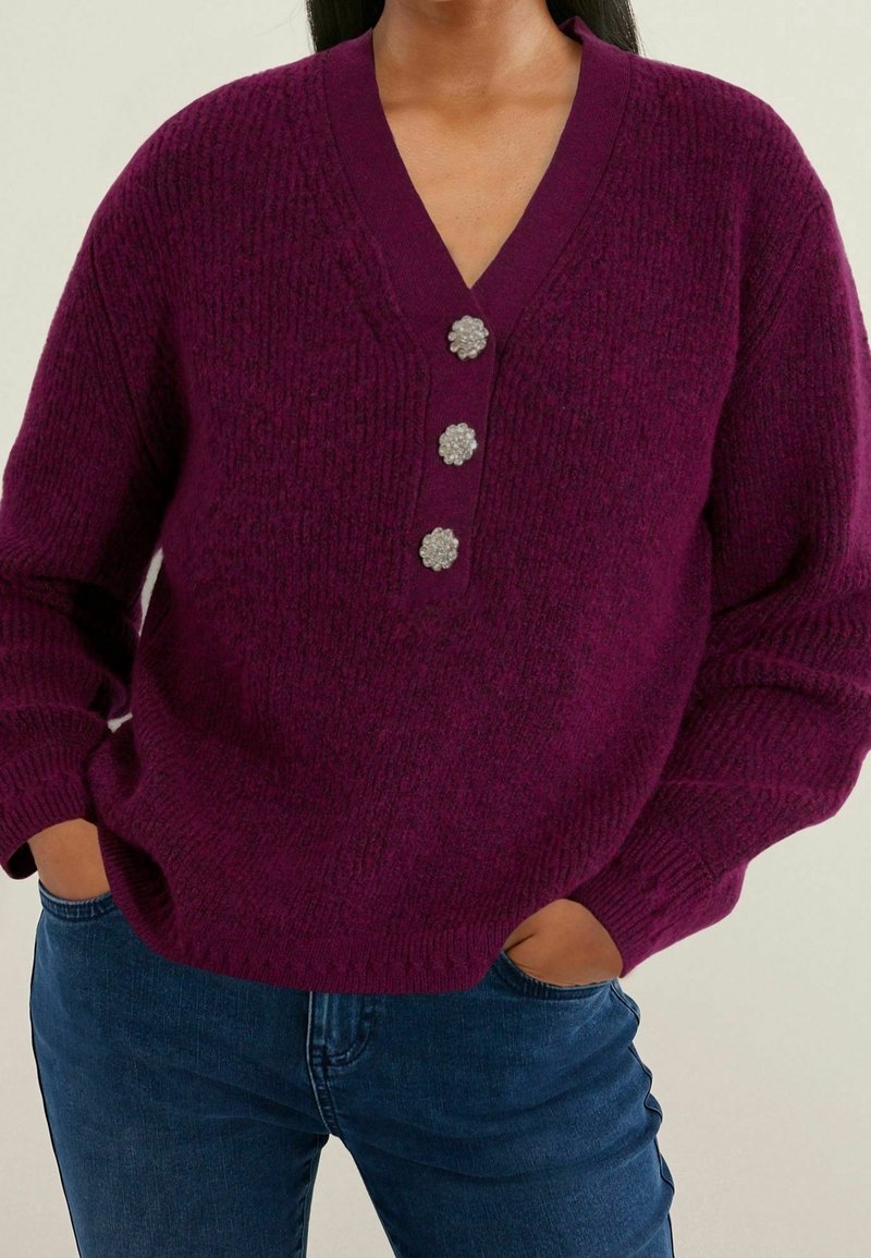 Suéter de punto morado con cuello en V y tres botones decorativos, con puños acanalados y un corte holgado. Combinado con vaqueros de mezclilla azul.