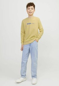 Felpa gialla con logo "JACK & JONES" blu, abbinata a jeans azzurri e sneaker bianche, in piedi contro uno sfondo semplice.
