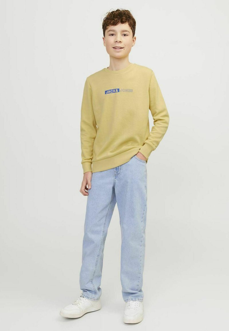 Felpa gialla con logo "JACK & JONES" blu, abbinata a jeans azzurri e sneaker bianche, in piedi contro uno sfondo semplice.