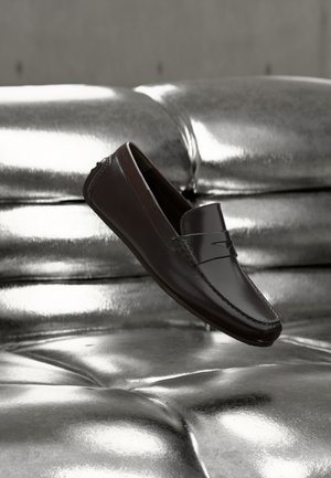 Mocassin en cuir marron foncé flottant au-dessus d'un canapé en cuir argenté brillant capitonné, mettant en valeur un design élégant et des détails de couture.