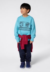 Sweatshirt turquesa com um logotipo "Champion" em azul escuro, suéter vermelho amarrado à cintura, calças de treino azul-marinho e ténis azuis.