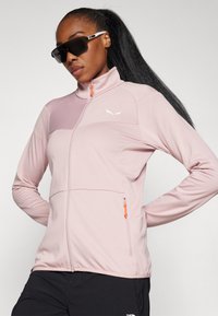 Lichtroze zip-up jack van gestructureerde stof, met hoge kraag en oranje ritsdetail. Beschikt over een strakke ontwerpen en een subtiele logo op de borst.
