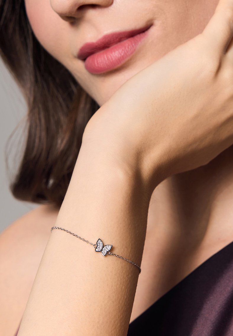 Srebrna bransoletka łańcuszkowa z zawieszką w kształcie motyla, ozdobiona małymi, przezroczystymi kamieniami. Ręka jest lekko opalona, o gładkiej fakturze.