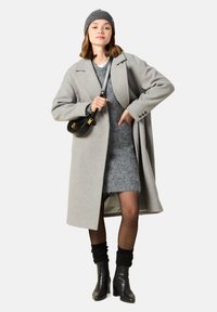 Grå ullkappa med snedkrage, kombinerad med en grå stickad klänning och svarta ankelstövlar. Tillbehör inkluderar en grå mössa och en svart handväska.