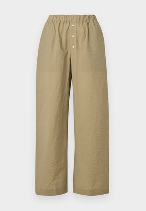 Mujer con cabello oscuro, camiseta blanca corta, pantalones anchos color oliva, zapatos negros, sosteniendo un bolso beige tejido, de pie frente a un fondo blanco liso.