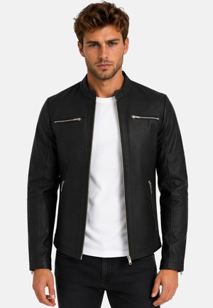 Hombre con pelo corto y rizado que lleva una chaqueta de cuero negra con cremallera sobre una camisa blanca y pantalones negros frente a un fondo liso.