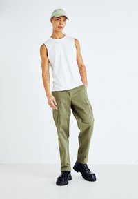 Top bianco senza maniche, pantaloni cargo verde oliva con tasche e stivali neri chunky con lacci. La modella indossa un berretto verde chiaro. Sfondo semplice.