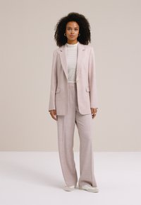 Moteris su banguotais plaukais, vilkinti šviesiai rožinį siūtą kostiumą, tekstūruotą baltą palaidinę ir baltus batus, stovinti priešais paprastą kreminės spalvos foną.