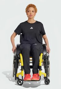 Donna con i capelli corti e ricci in un completo Adidas nero, seduta su una sedia a rotelle gialla e indossando sneaker arancioni brillanti, rivolta in avanti contro uno sfondo bianco.