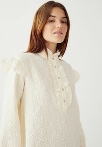 Kreemimaitseline nööbitav pluus, millel on volangiga krae ja varrukad, tekstuuriga kangas ning ees olevad nööbid. Lihtne, elegantne disain pehmete aksentidega.