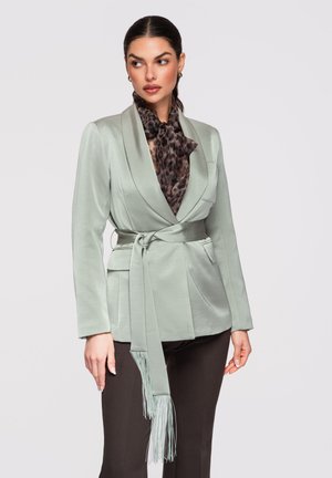 Femme portant un blazer gris clair ceinturé sur un chemisier à imprimé léopard, associé à un pantalon foncé, regardant légèrement sur le côté.