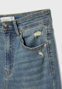 Section avant d'un jean en denim bleu avec bouton, passants pour ceinture, deux patchs usés et une petite poche avant montrant des détails de tissu usé.