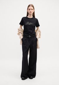 Sort bomulds t-shirt med et glitrende design, parret med højhalsede sorte bukser og en beige knap-skjorte, der har et bælte med en cirkulær spænde.