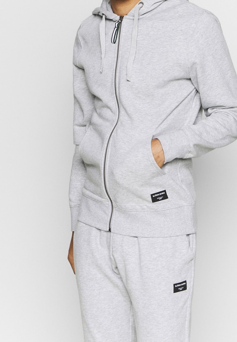 Sweat à capuche gris à fermeture éclair fabriqué en tissu doux avec une poche avant et une capuche à cordon. Associé à un pantalon de survêtement assorti avec des écussons logo.