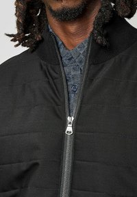 Veste de bombardier noire matelassée avec fermeture éclair à l'avant, col côtelé et une chemise à carreaux bleue visible en dessous. Présente un détail de fermeture éclair argenté.