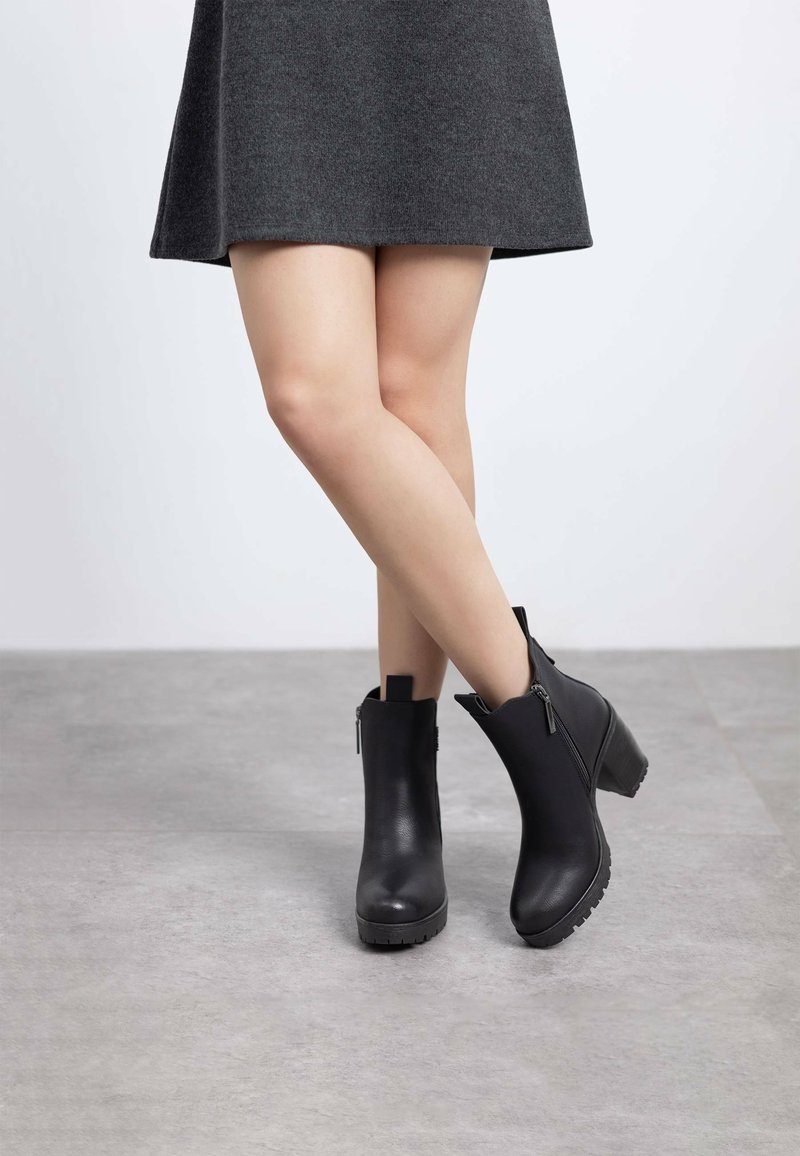 Botas de cuero negras hasta el tobillo con un tacón grueso, con cremallera lateral y suela texturizada, llevadas con una falda gris sobre una superficie lisa.