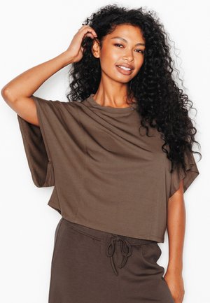 Femme aux longs cheveux bouclés portant un haut marron ample et un pantalon, souriante, une main touchant ses cheveux.