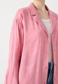 Personne portant une chemise rose ample à boutons avec des motifs subtils de feuilles de palmier sur un débardeur blanc et un jean bleu, raccourcie aux épaules et à la taille.