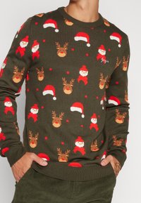 Grön stickad tröja med ett festligt mönster av jultomte, renar och julhattar i röd, brun och vit. Ribbstickade ärmslut och fåll.